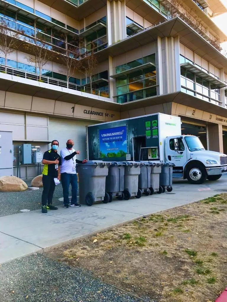 Two men are standing outside a large building, with garbage cans positioned in the foreground.