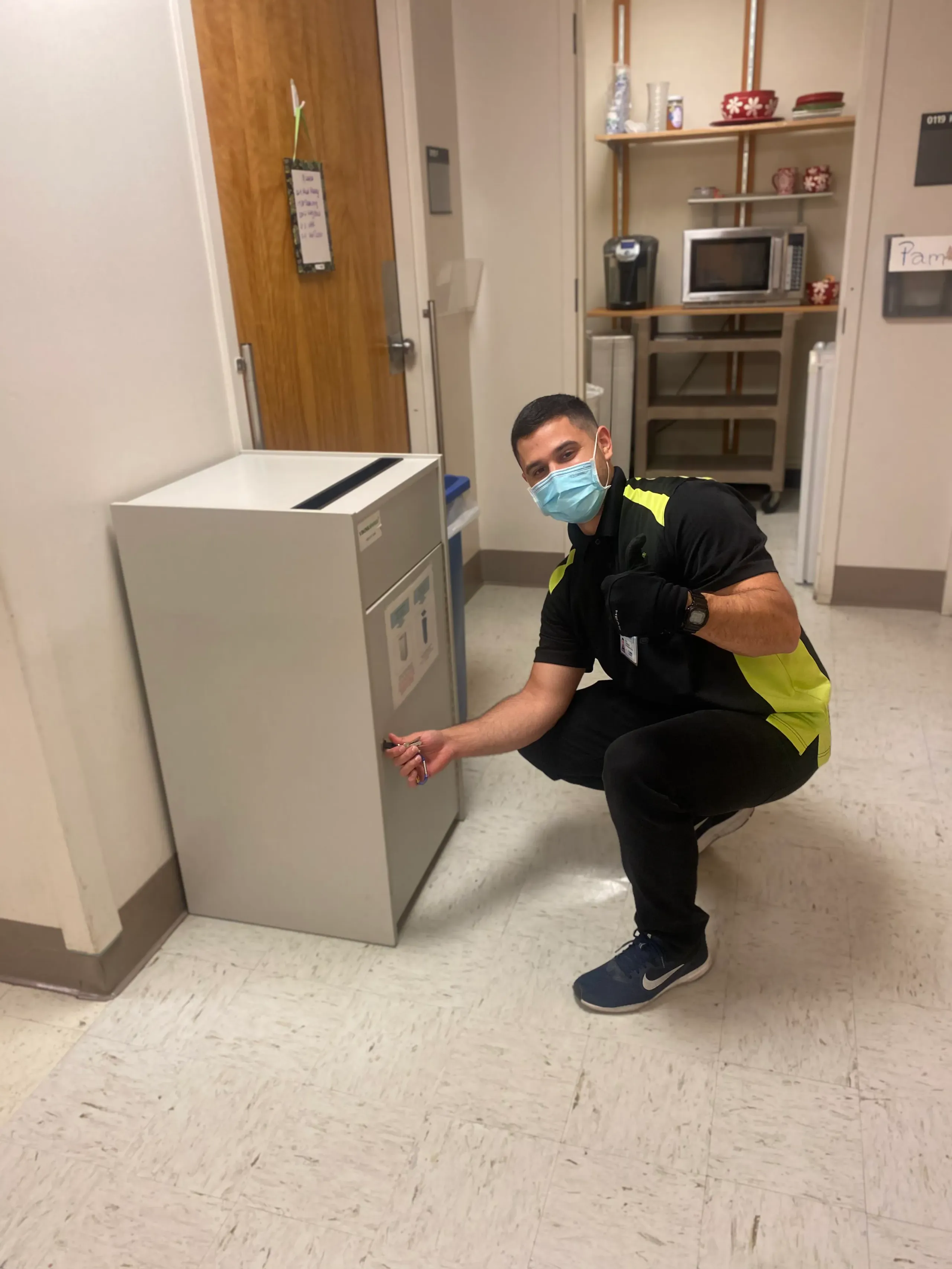 A man in protective gear kneels next to a refrigerator, likely examining its interior or performing a service.