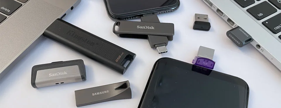 An overhead view of various USB flash drives and adapters, some plugged into a smartphone and laptops.