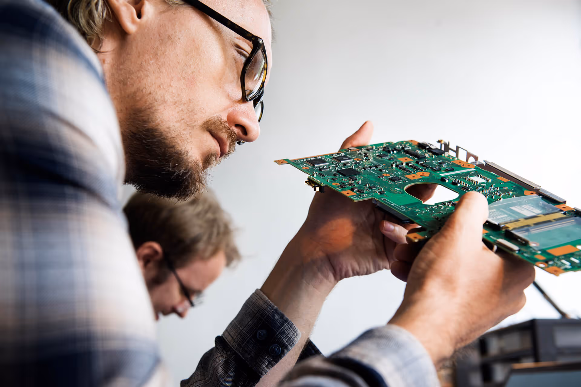 A close-up side view of a technician wearing glasses intently inspecting a green computer motherboard.