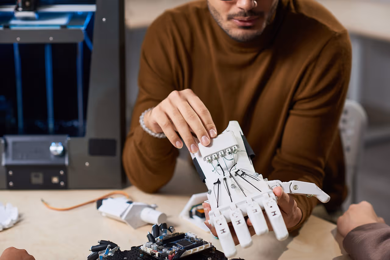 Engineer examining a 3D-printed robotic hand prototype in a lab with a 3D printer in the background.