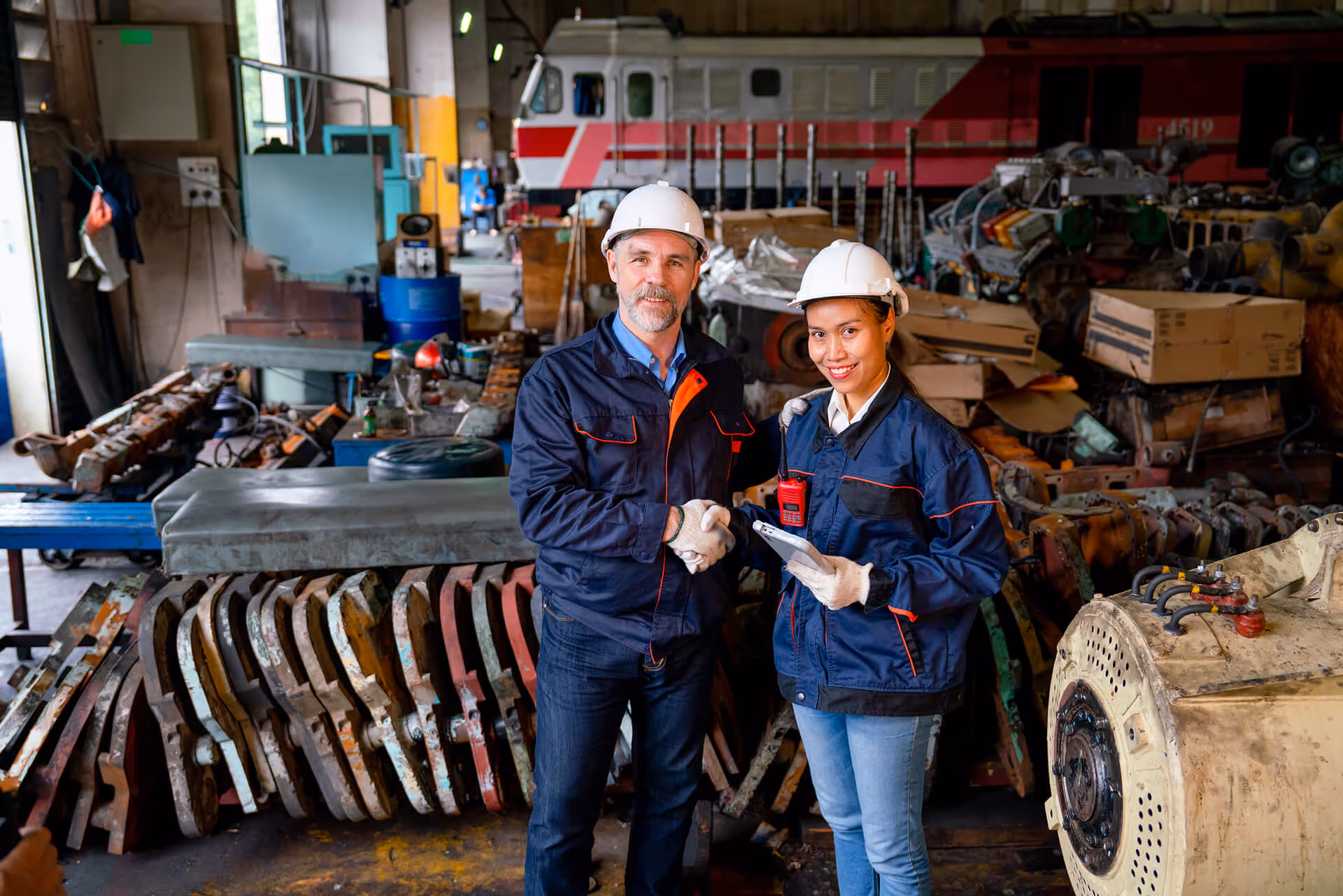 Two industrial engineers in hard hats and uniforms shaking hands in a train maintenance workshop.
